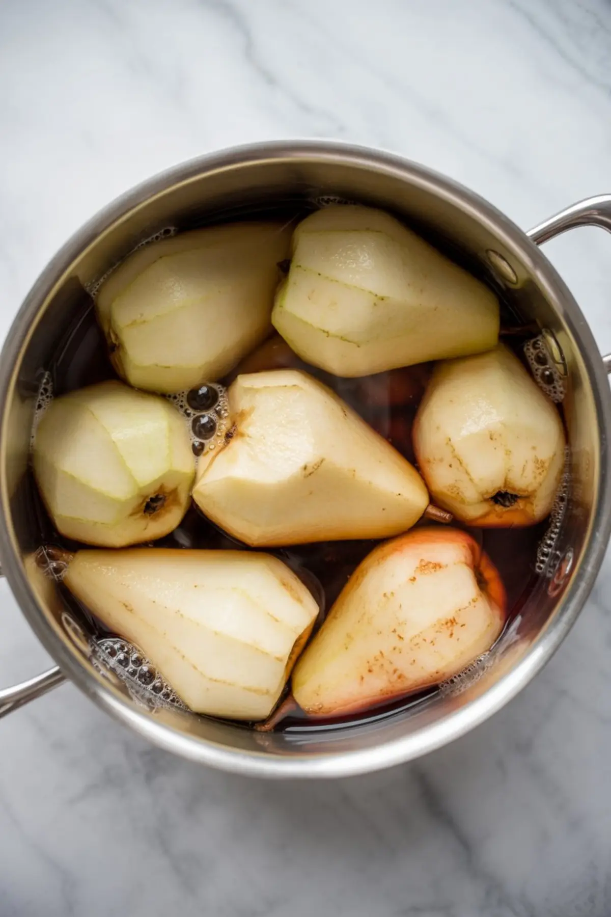 Peeled pears submerged in a pot of spiced red wine poaching liquid during the cooking process for poached pears.

