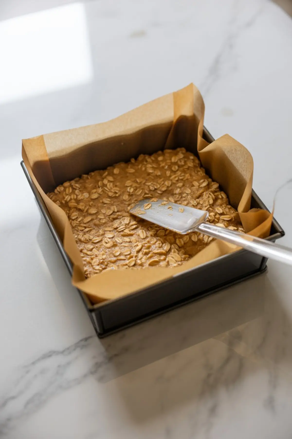 Square baking pan lined with parchment paper filled with an even layer of oat and peanut butter mixture being smoothed with a metal spatula on a marble countertop.