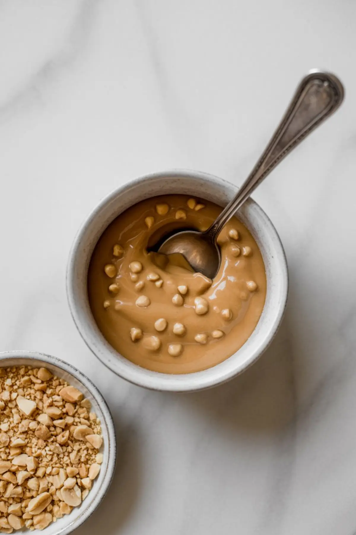Small ceramic bowl of creamy peanut butter with peanut butter chips and a spoon, placed beside a bowl of chopped peanuts on a white marble surface.