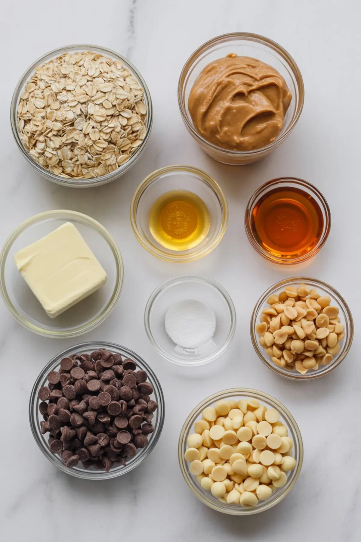 Overhead view of measured ingredients in glass bowls, including rolled oats, peanut butter, butter, honey, vanilla extract, salt, white chocolate chips, milk chocolate chips, and peanuts on a marble surface.