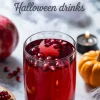 Glass of pomegranate juice with floating seeds on a Halloween-themed setup, surrounded by a whole pomegranate, a small pumpkin, and a lit tealight candle on a marble surface.