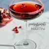 A pomegranate martini garnished with floating pomegranate seeds and a pink sugar rim, served in a coupe glass on a sunlit marble surface with a halved pomegranate in the background.