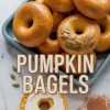 Tray of glossy pumpkin bagels, some topped with pumpkin seeds, with a sliced bagel spread with cream cheese. Text overlay reads “Pumpkin Bagels.”