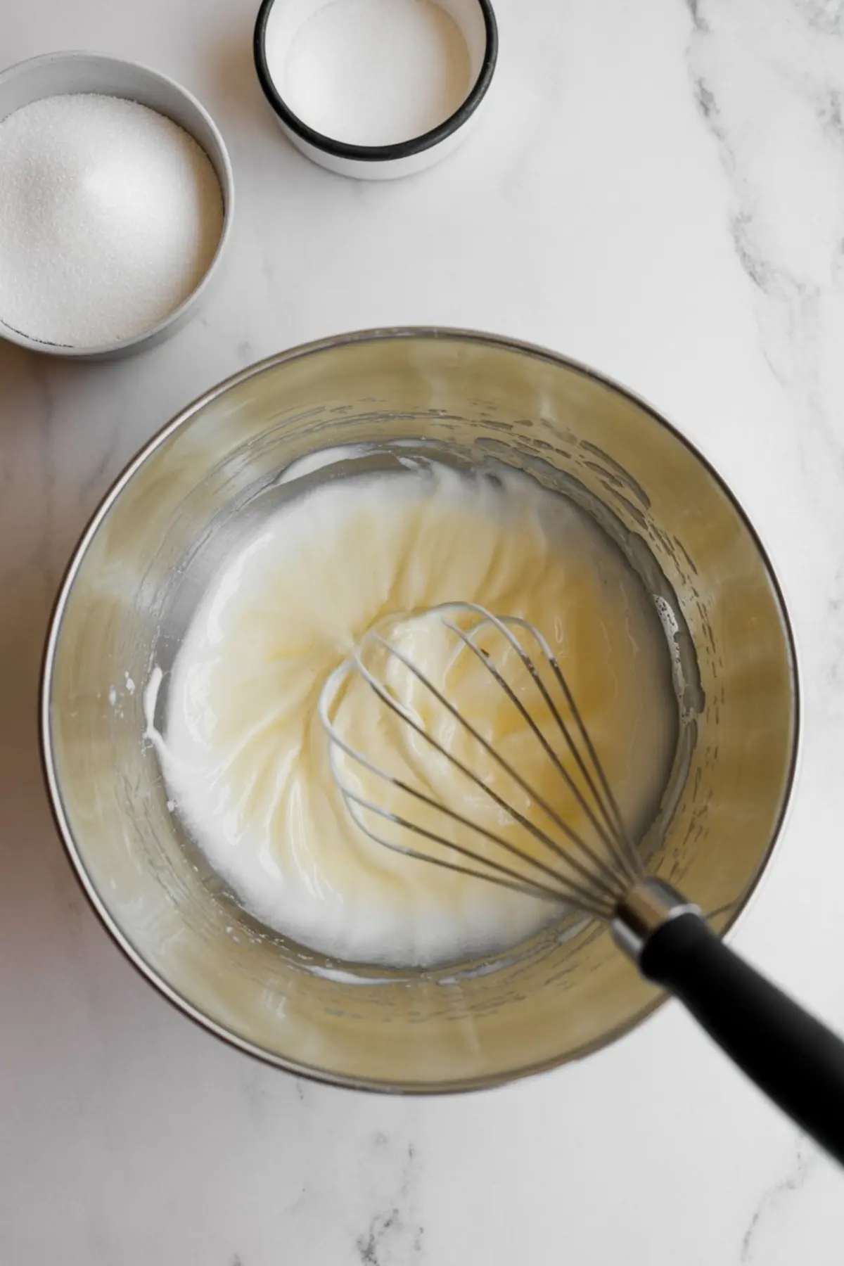 Stainless steel mixing bowl with egg whites being whisked to soft peaks, surrounded by small bowls of granulated sugar on a white marble counter.