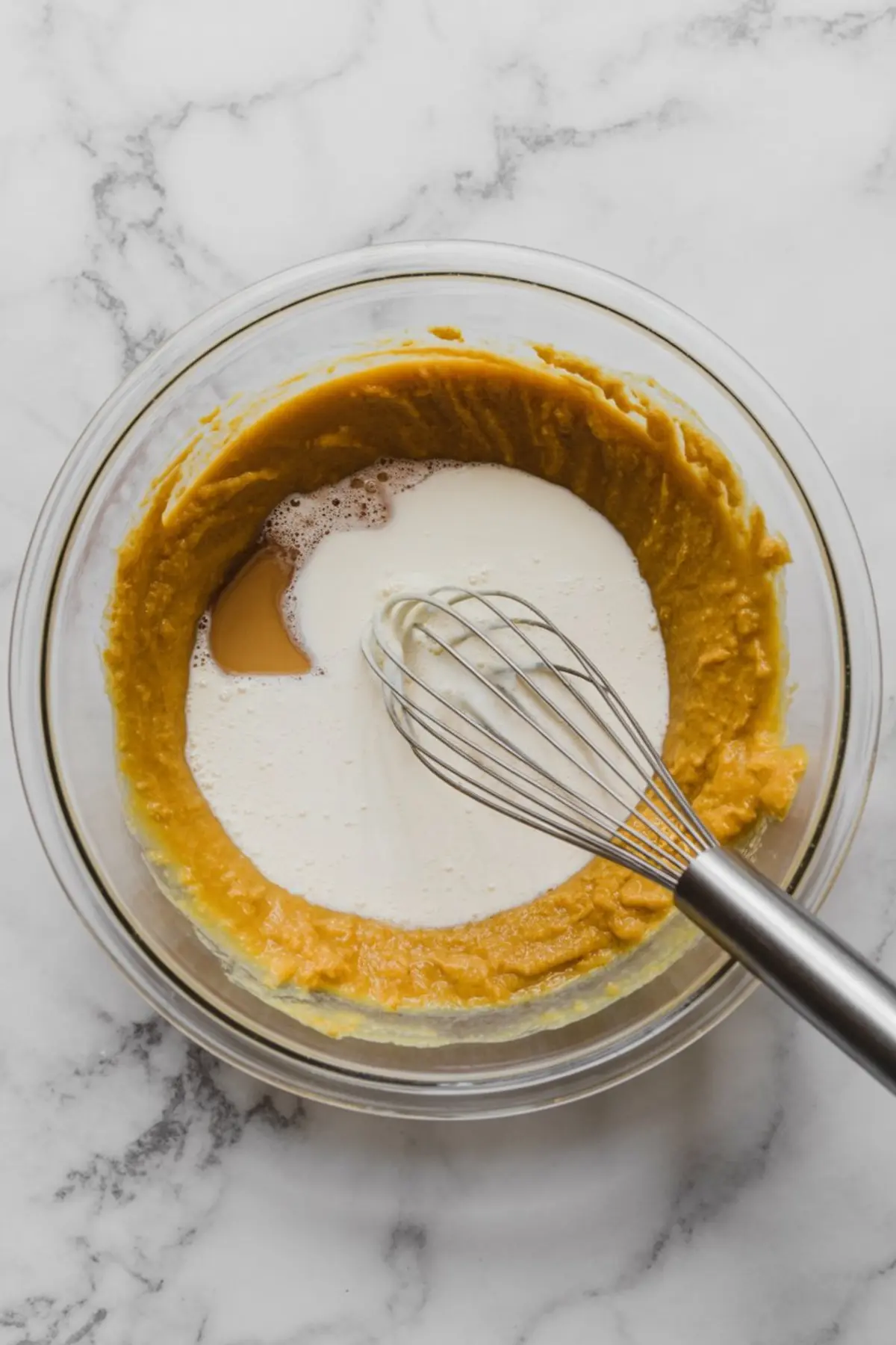Glass bowl filled with pumpkin puree being mixed with cream and vanilla using a metal whisk on a marble surface, showing the process of making pumpkin ice cream base.