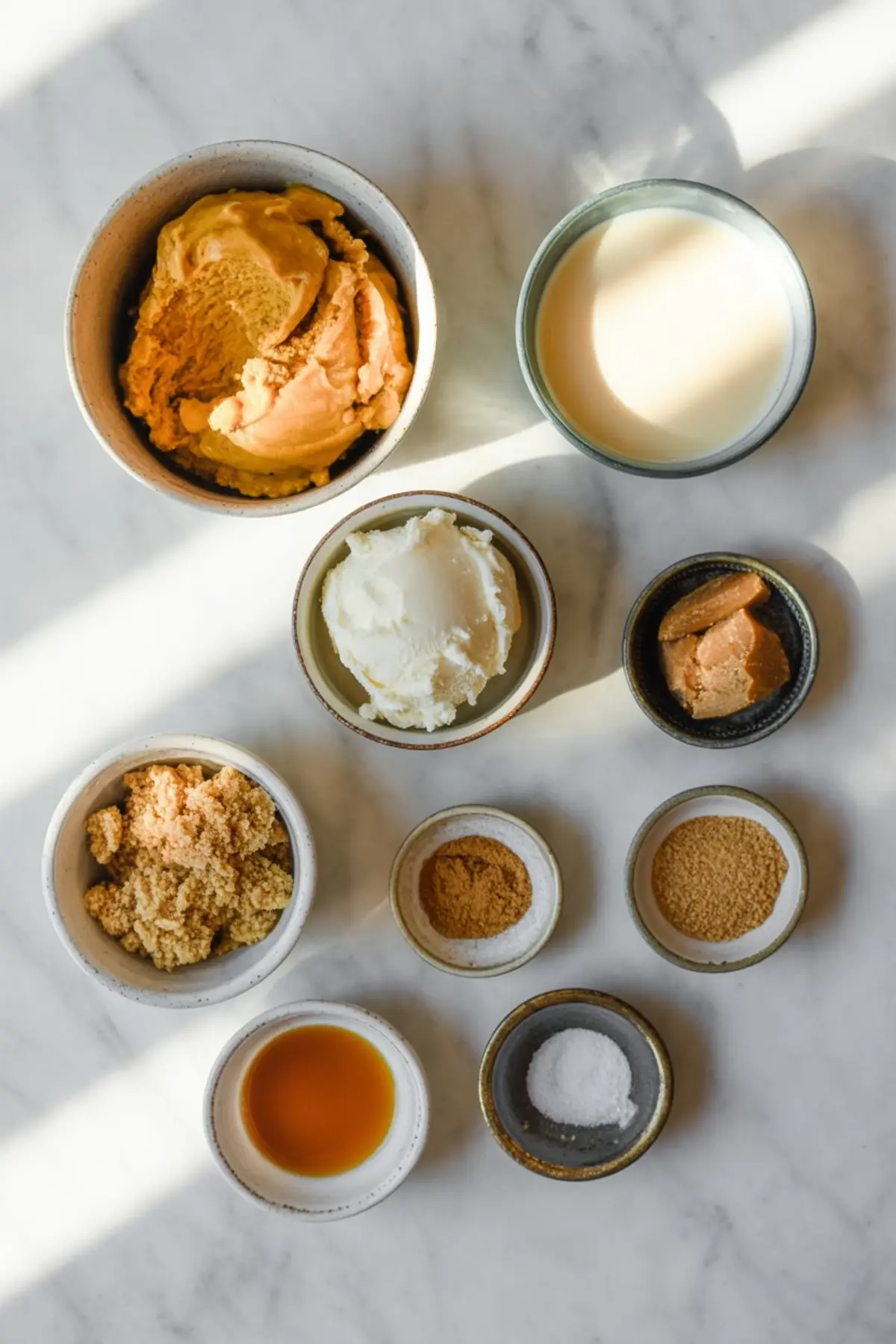 Flat lay of individual pumpkin ice cream ingredients in ceramic bowls, including pumpkin puree, brown sugar, heavy cream, vanilla extract, cream cheese, spices, and salt on a marble countertop in soft sunlight.
