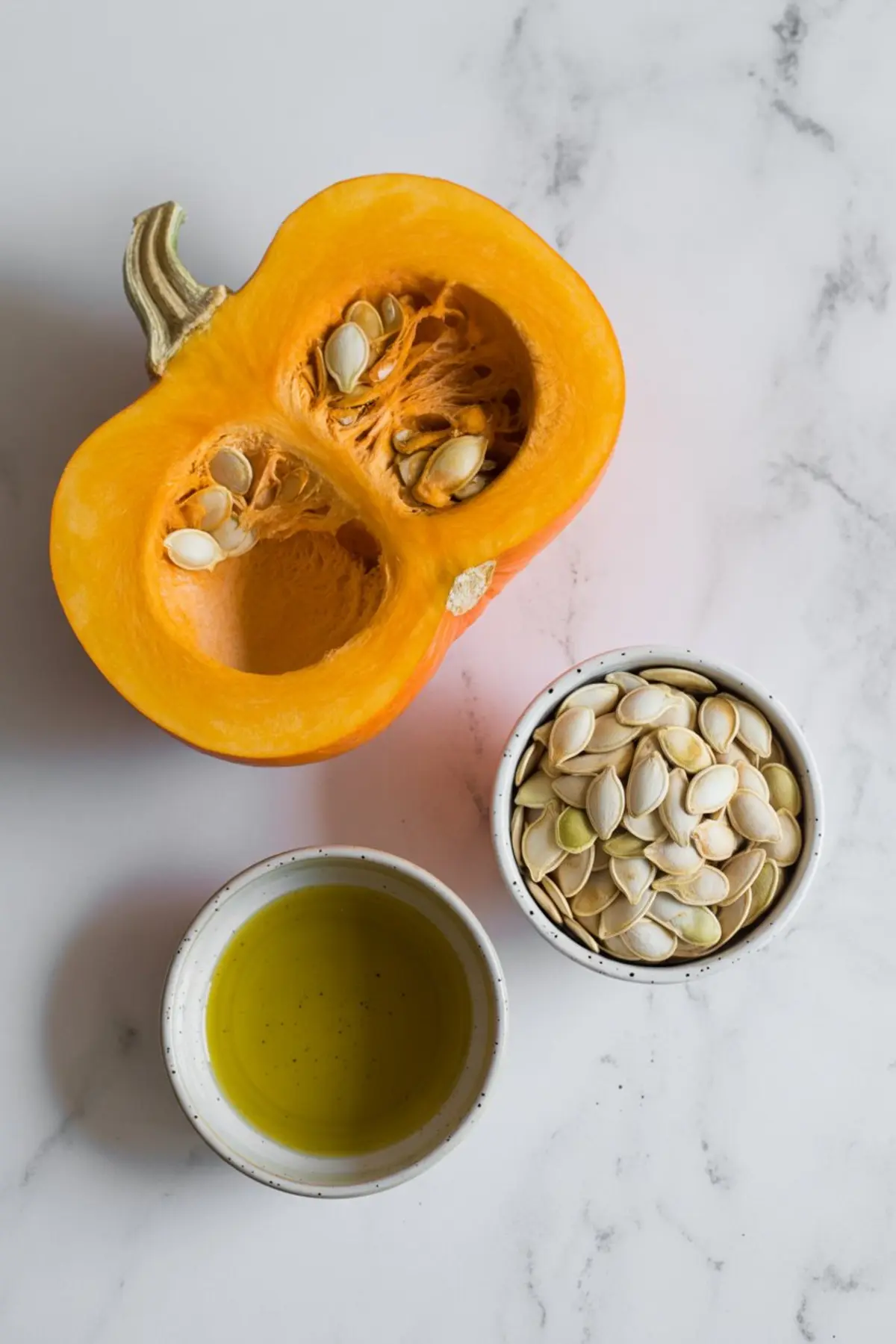 Fresh pumpkin half with seeds inside, placed on a marble surface beside a small bowl of raw pumpkin seeds and a bowl of golden pumpkin seed oil.
