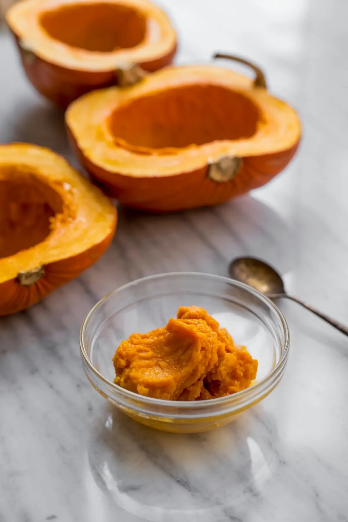 Glass bowl filled with smooth homemade pumpkin purée on a marble counter, with roasted pumpkin halves and a spoon in the background.
