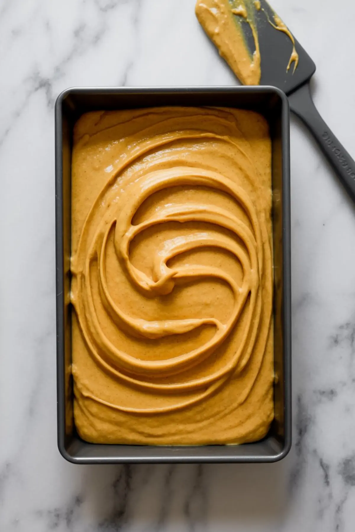 Pumpkin batter spread evenly in a rectangular metal baking pan with a spatula placed nearby on a marble background, showing smooth, creamy texture ready for baking.
