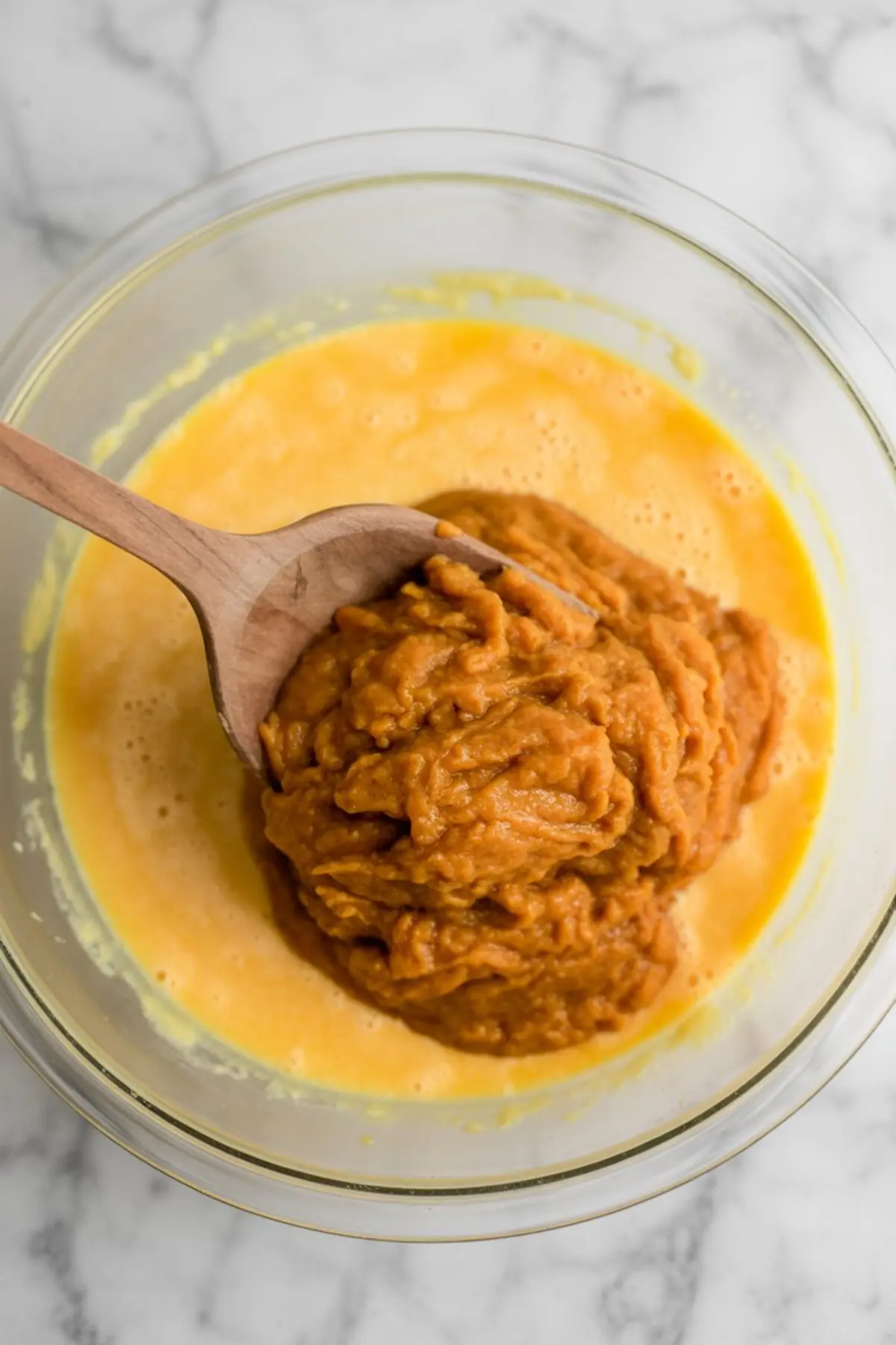 Glass bowl with beaten egg mixture and a wooden spoon scooping thick pumpkin puree into the wet ingredients, highlighting the base for a spiced pumpkin cake batter.
