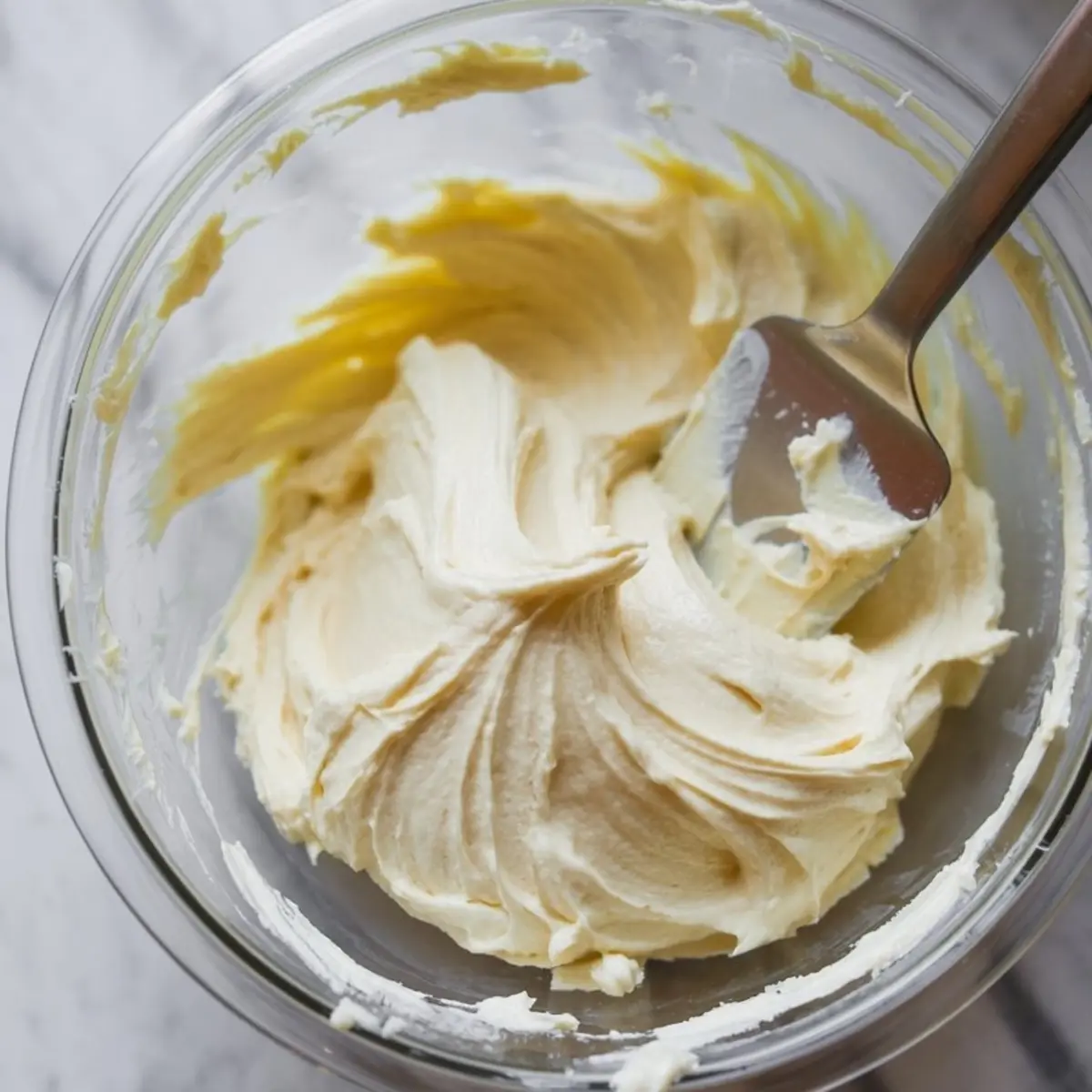 Glass bowl of smooth vanilla frosting with a spatula, showcasing the final creamy texture for topping pumpkin cake or bars.

