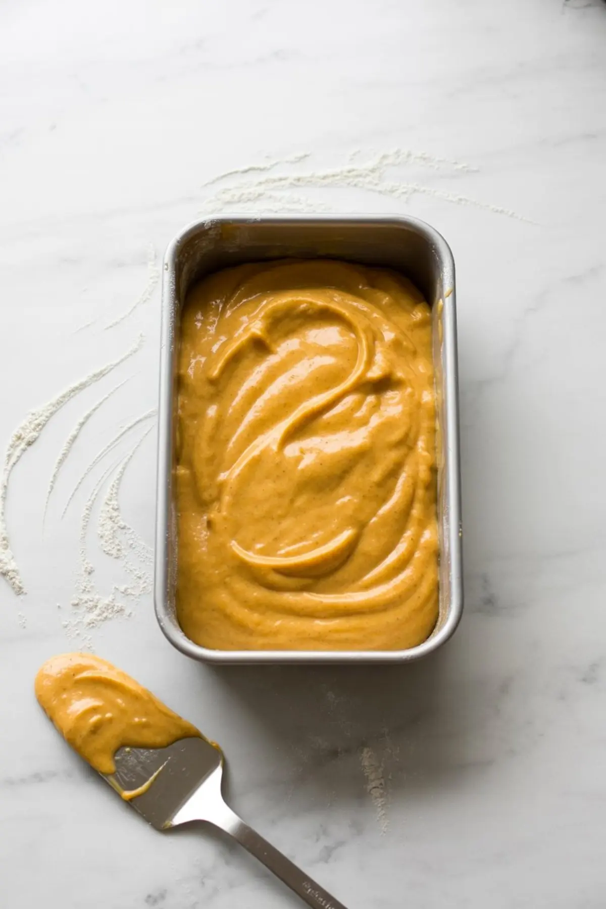 Rectangular baking pan filled with smooth pumpkin cake batter, with a spatula resting beside it on a floured marble countertop.