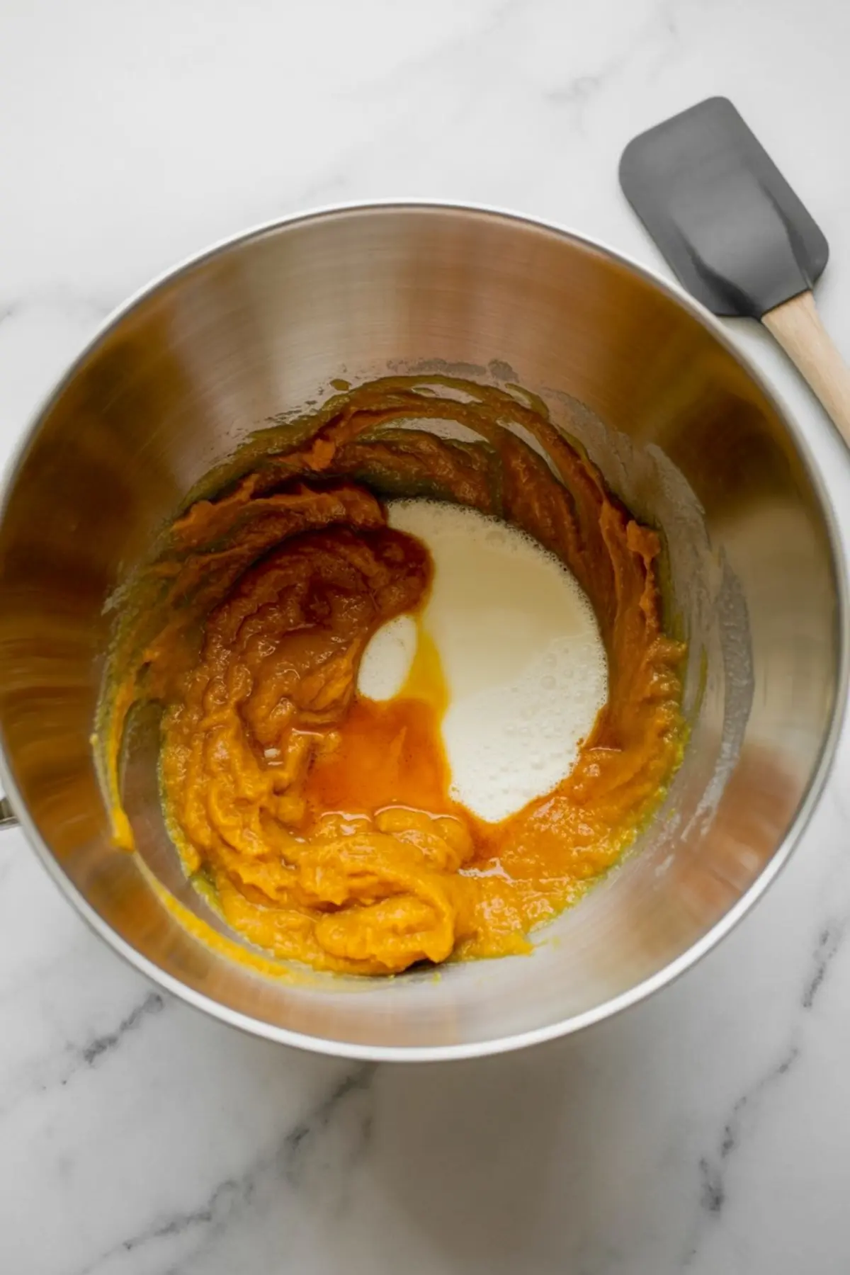 Mixing bowl with pumpkin puree, sugar, and wet ingredients being combined for a pumpkin cake batter on a marble surface with a silicone spatula nearby.