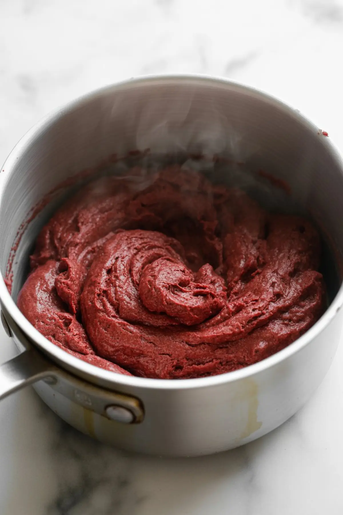 Freshly cooked red velvet churro dough in a metal saucepan, with steam rising from the thick, velvety red mixture. The swirled texture emphasizes its rich and smooth consistency.