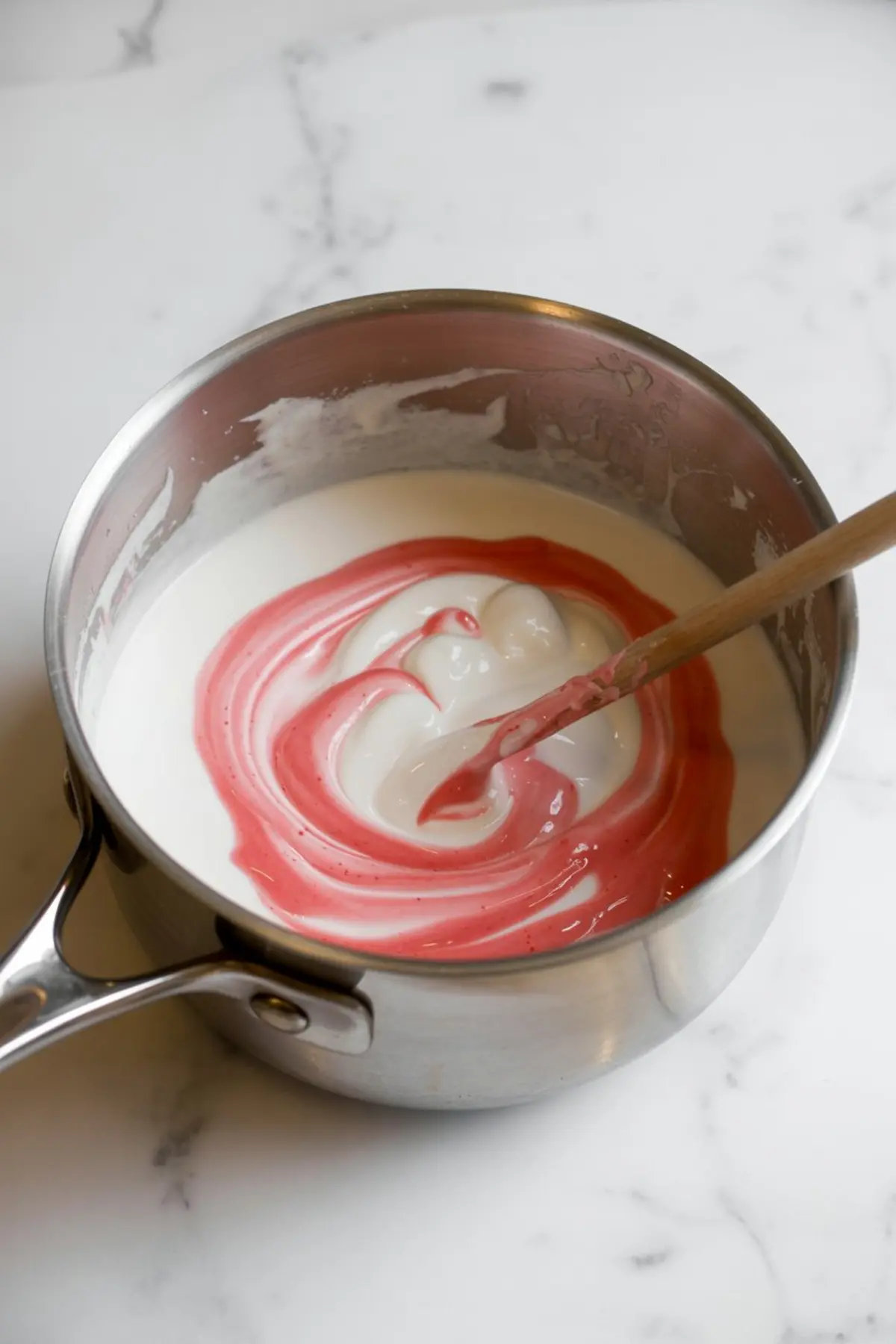 Pink food coloring being swirled into a pot of melted white marshmallow mixture with a wooden spoon on a white marble surface.