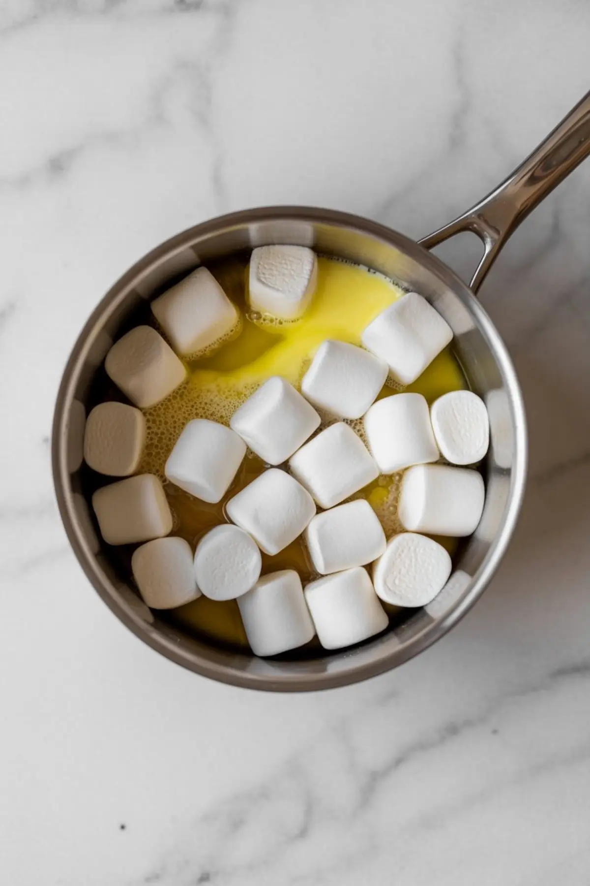 Whole marshmallows melting into a pool of melted butter inside a stainless steel saucepan on a marble counter.