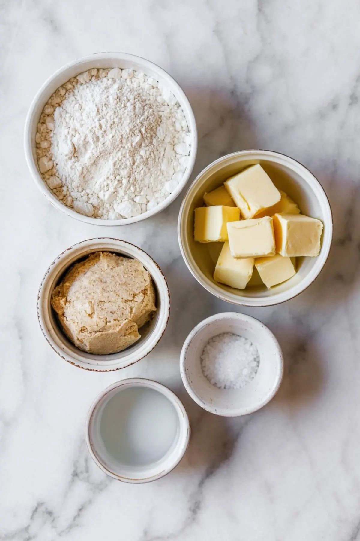 Overhead view of baking ingredients in ceramic bowls on a marble surface, including all-purpose flour, cubed butter, brown sugar, coarse salt, and milk.