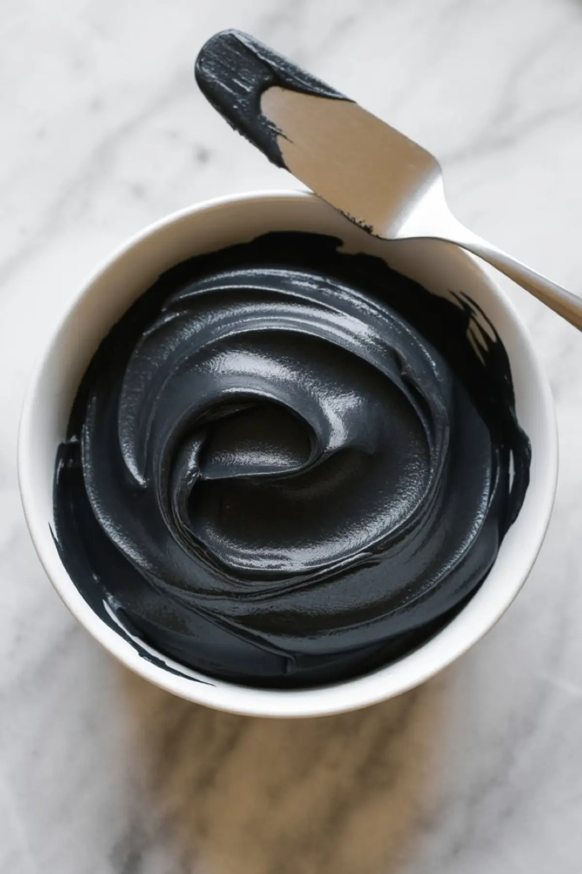 Bowl of glossy black frosting with a metal spatula coated in icing, placed on a light marble surface.