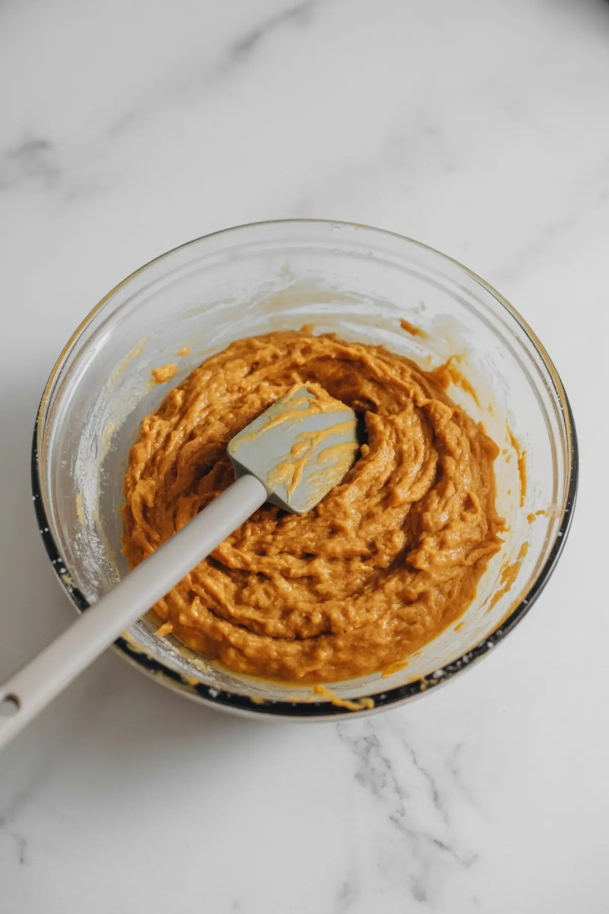 Glass mixing bowl filled with sweet potato muffin batter, stirred with a grey silicone spatula on a white marble countertop.