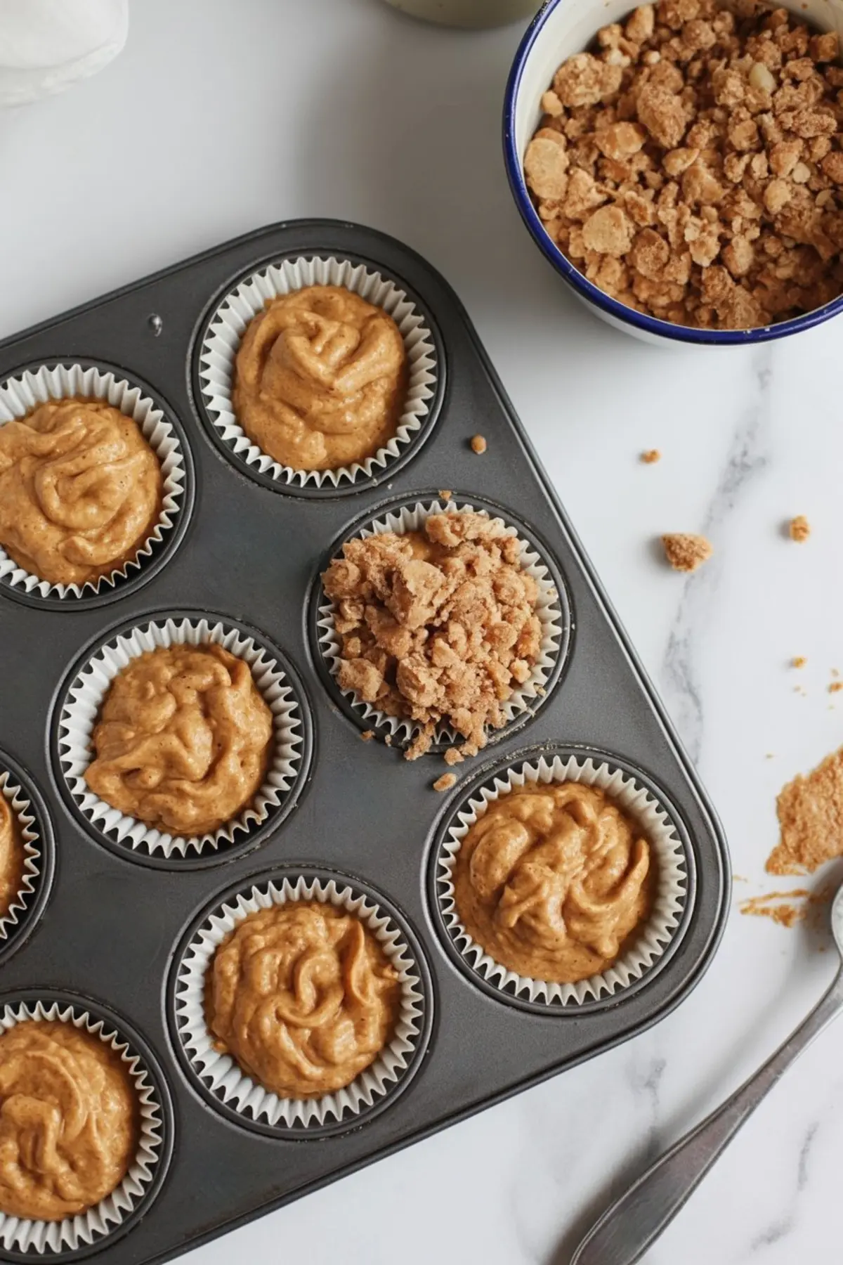 Muffin tin with sweet potato batter in paper liners, one topped with crumbly streusel, surrounded by a bowl of topping and spoon on a white surface.