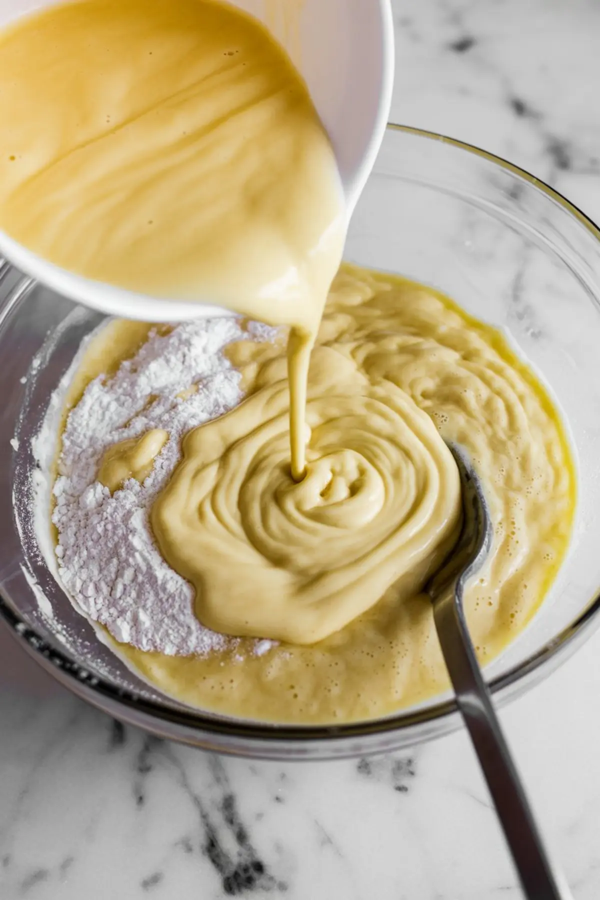 Thick sweet potato pancake batter being poured from a white bowl into a glass mixing bowl with flour on a marble countertop.