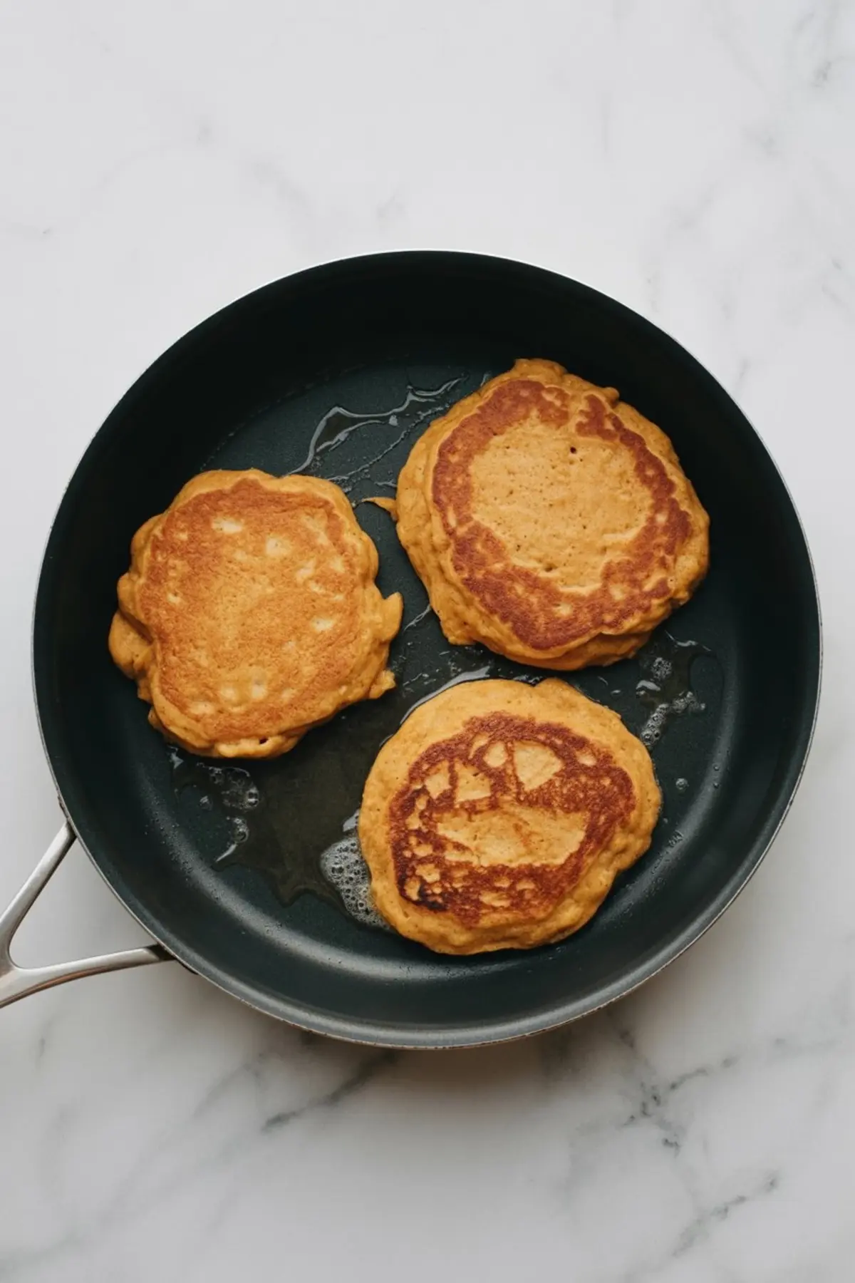 Three golden brown sweet potato pancakes sizzling in a non-stick skillet with a light layer of oil.