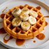Golden sweet potato waffle topped with banana slices and generously drizzled with maple syrup, served on a white plate with syrup accents.