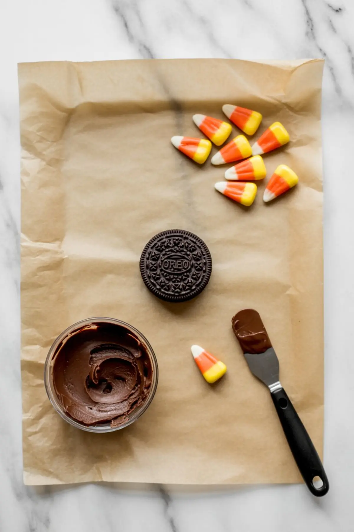 Flat lay of Oreo cookie, chocolate frosting, candy corn, and an offset spatula on parchment paper, showing ingredients and tools for making Thanksgiving turkey treats.
