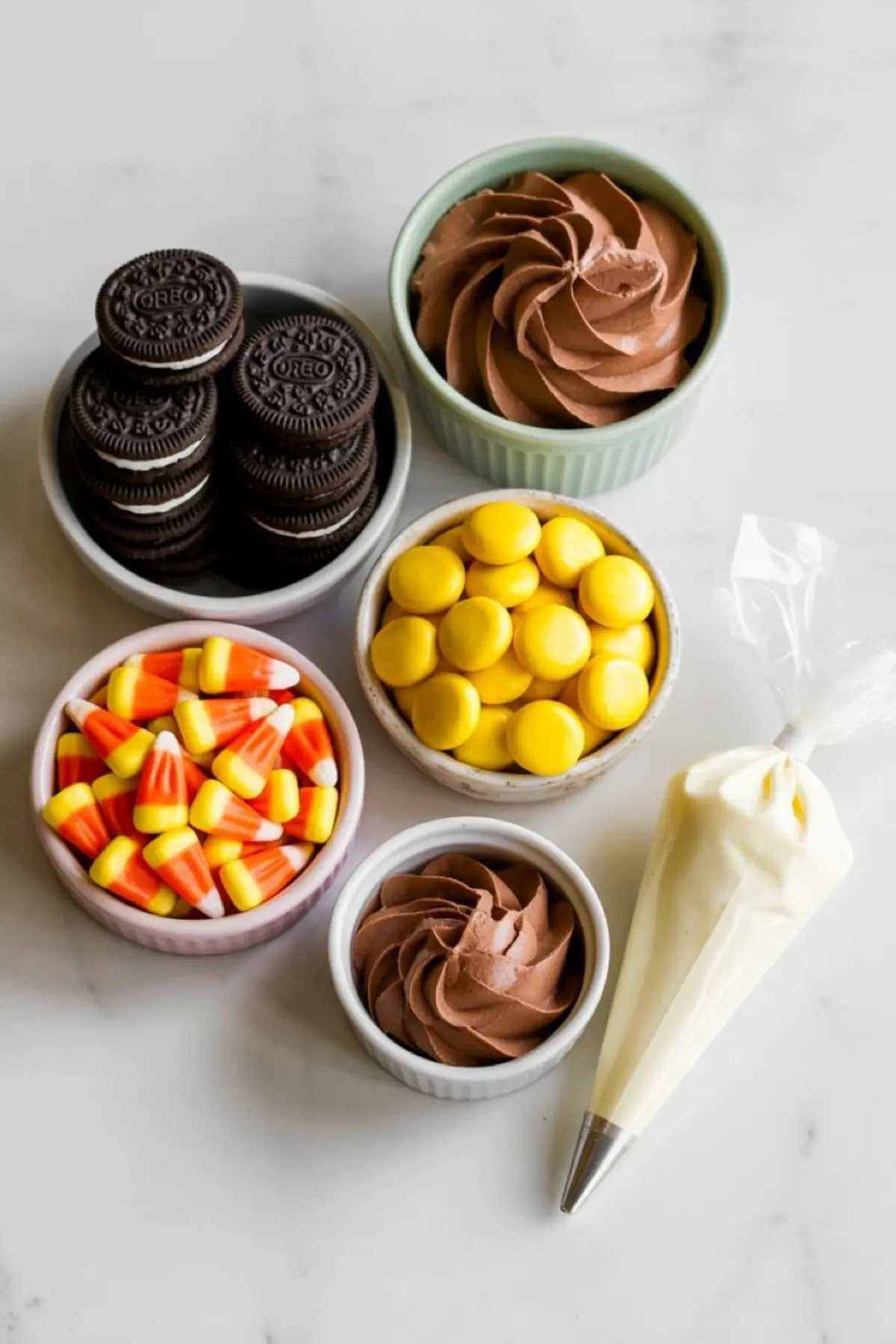 Assortment of Thanksgiving treat ingredients in small bowls, including Oreo cookies, candy corn, yellow chocolate candies, chocolate frosting, and a piping bag filled with white icing, arranged on a white surface.
