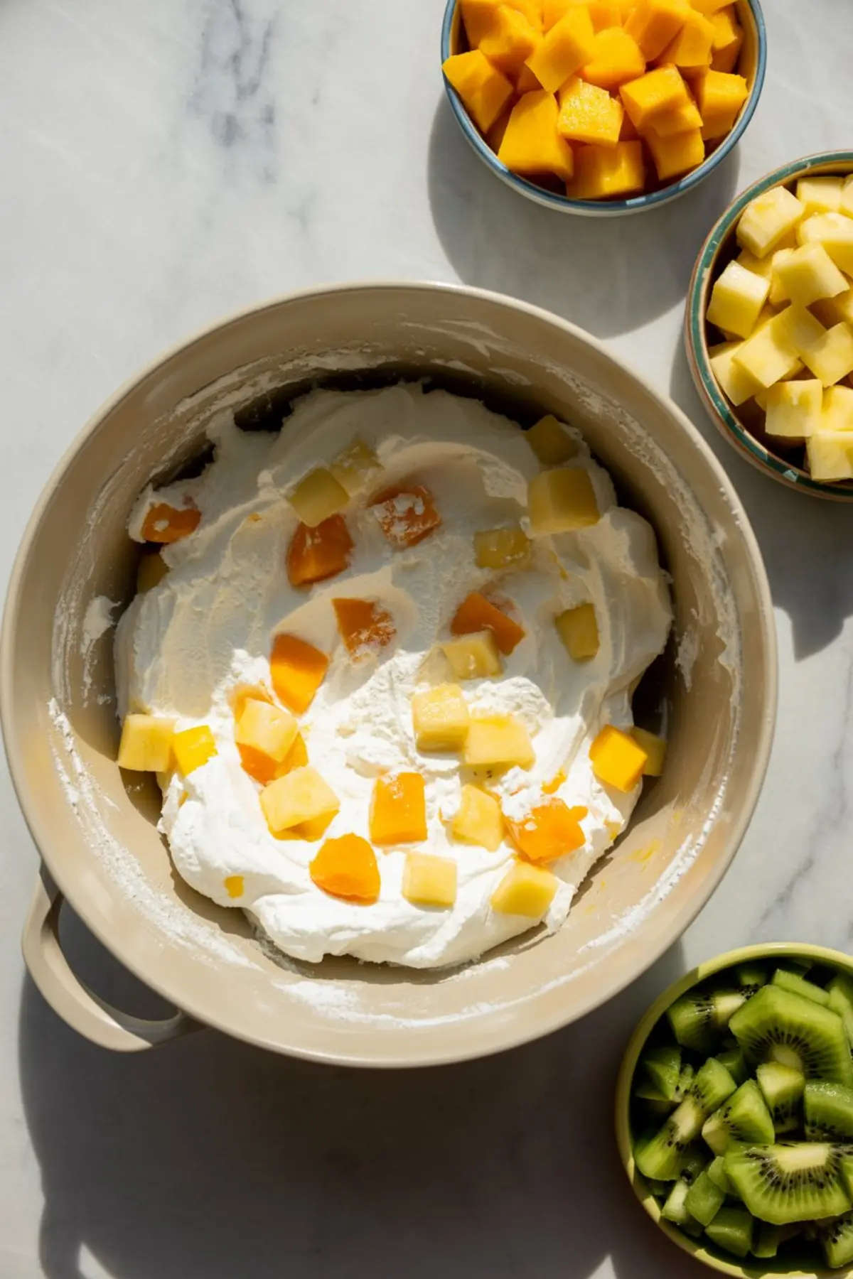 Mixing bowl of whipped cream topped with chunks of mango and pineapple, surrounded by bowls of diced kiwi, pineapple, and mango.