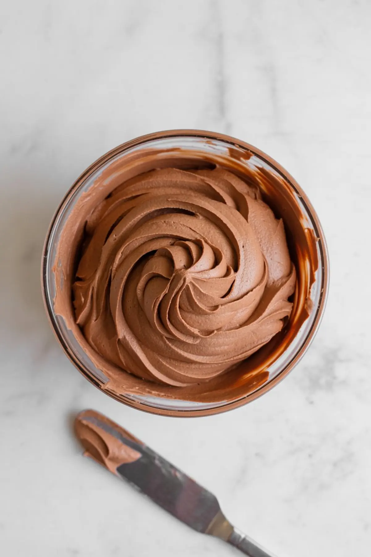 Bowl of creamy chocolate buttercream frosting with swirled texture, ready for cupcake decorating, placed on a marble countertop with an offset spatula beside it.
