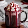 Steaming hot chocolate served in a black glass mug with a rim coated in red sugar crystals, topped with whipped cream and red syrup, dripping dramatically for a gothic Halloween drink display.