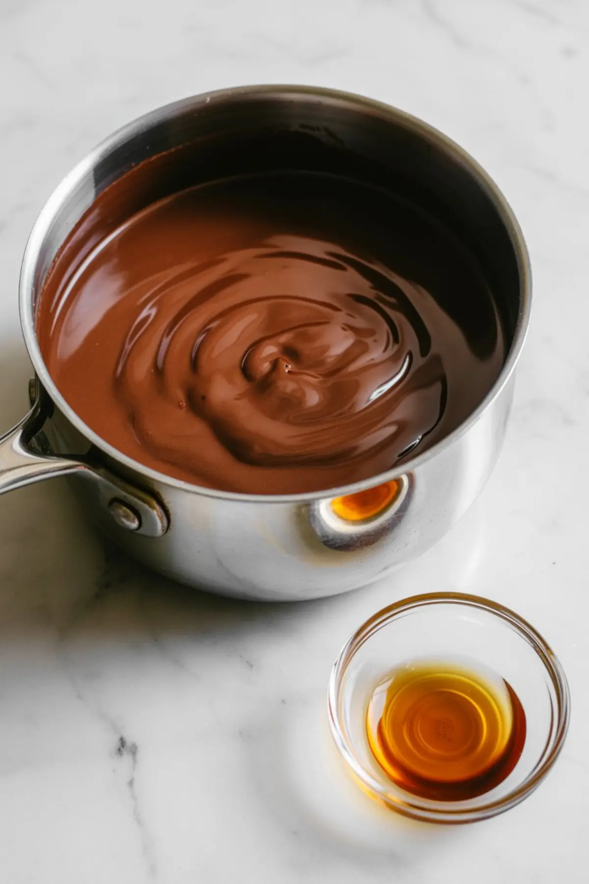 Creamy melted chocolate swirled in a stainless steel saucepan, with a small bowl of vanilla extract placed beside it on a marble countertop during hot chocolate preparation.

