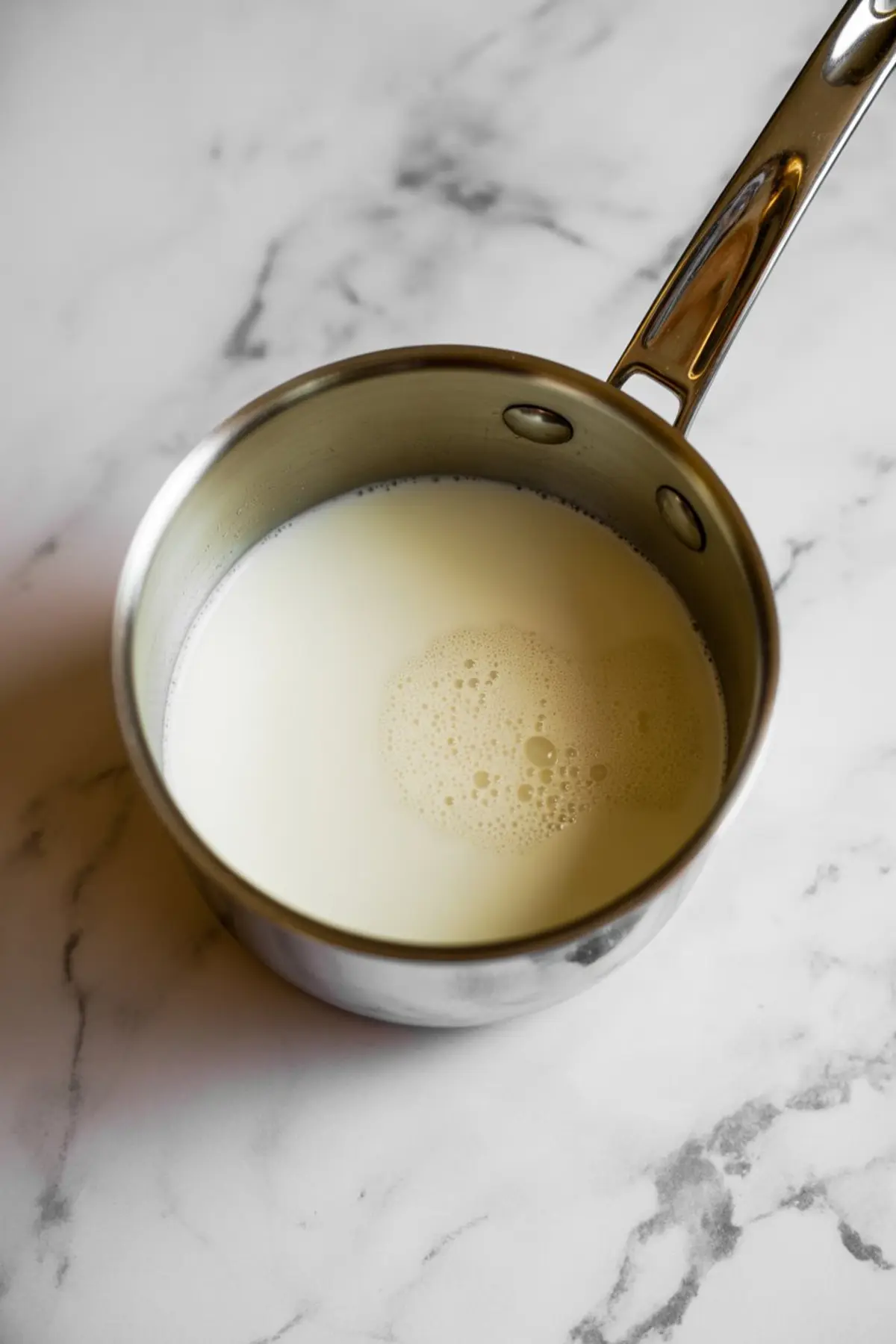 Warm milk heating in a stainless steel saucepan on a marble surface, shown as part of the process for making rich homemade hot chocolate.

