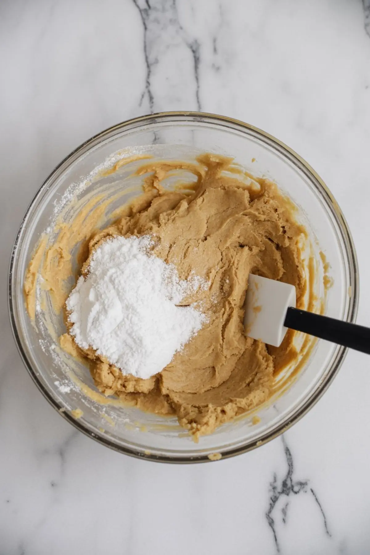 Glass mixing bowl with thick cookie dough, featuring a mound of white powdered sugar and a spatula, on a white marble countertop.