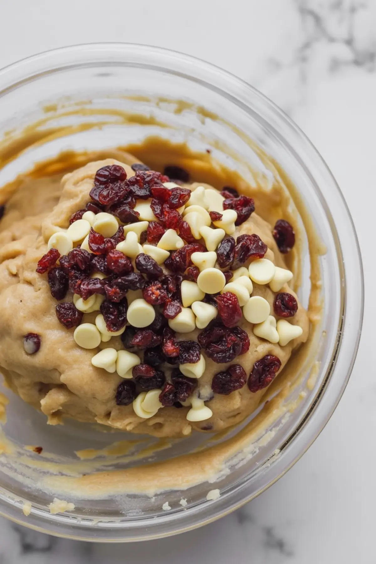 Cookie dough in a clear bowl topped with white chocolate chips and dried cranberries, ready for mixing.