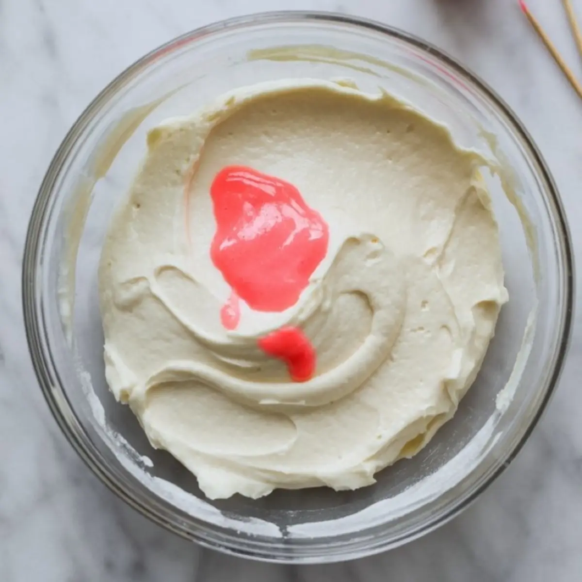 Vanilla frosting in a glass bowl with bright pink food coloring drizzled on top before blending.