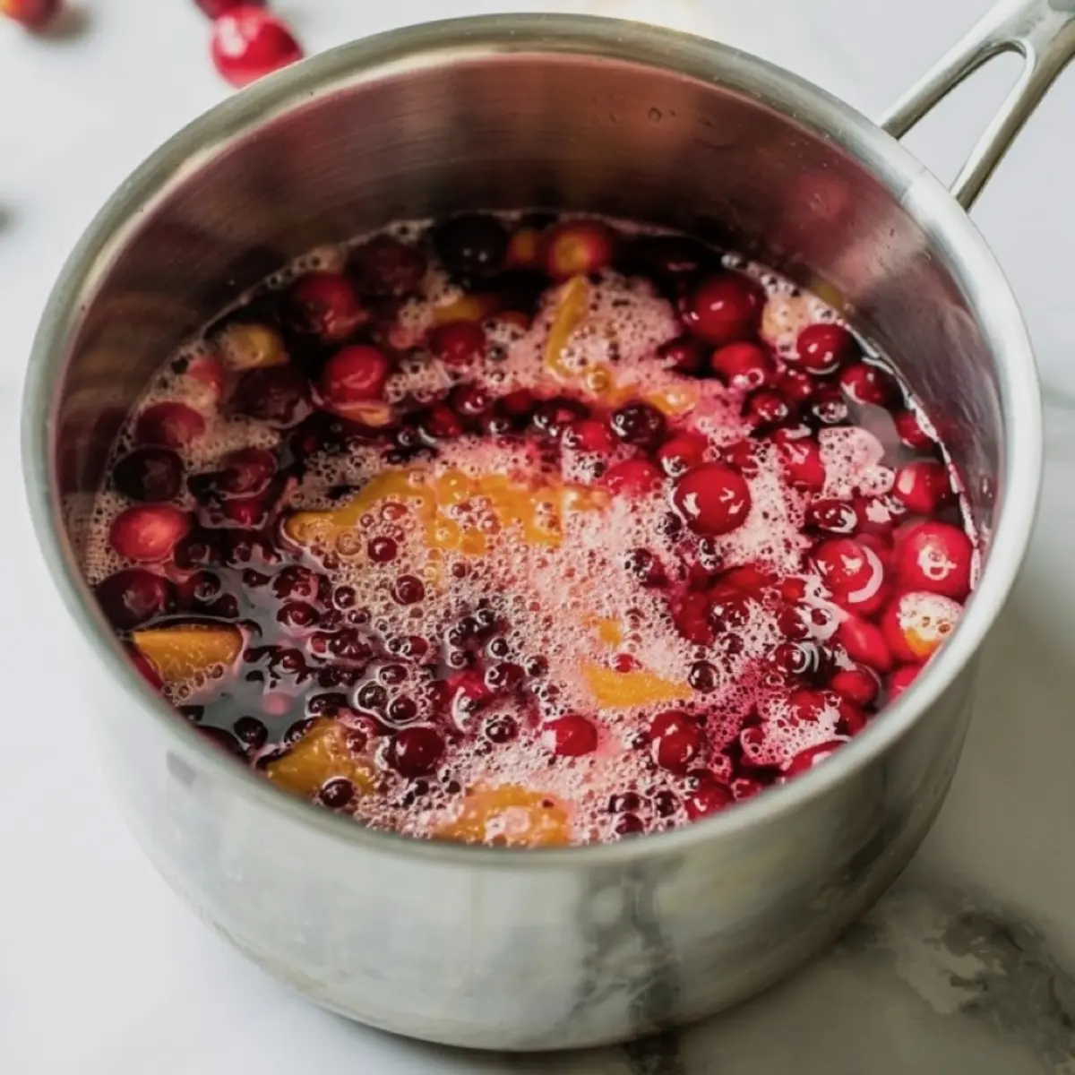 Close-up of a saucepan filled with simmering cranberries, orange slices, and juice, showing the process of making homemade fruit compote or sauce for baking.
