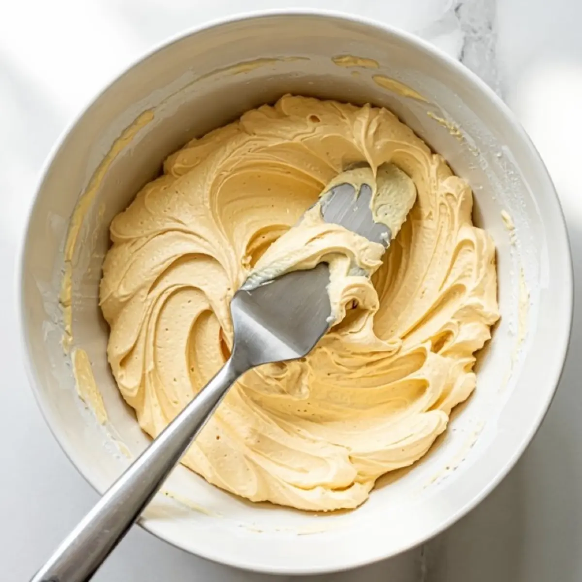Creamy cake batter swirled in a mixing bowl with a metal spatula, showcasing the smooth texture and rich consistency of homemade cake mixture.
