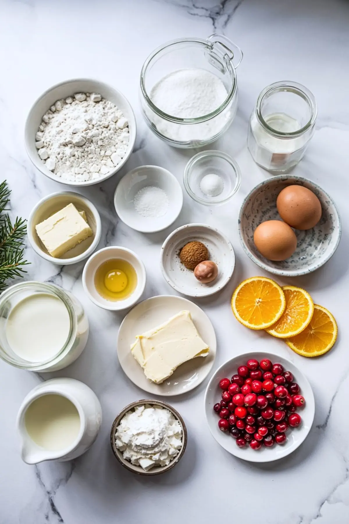 Flat lay of baking ingredients including flour, sugar, butter, eggs, cranberries, orange slices, milk, vanilla, nutmeg, and baking powder, arranged on a marble countertop.
