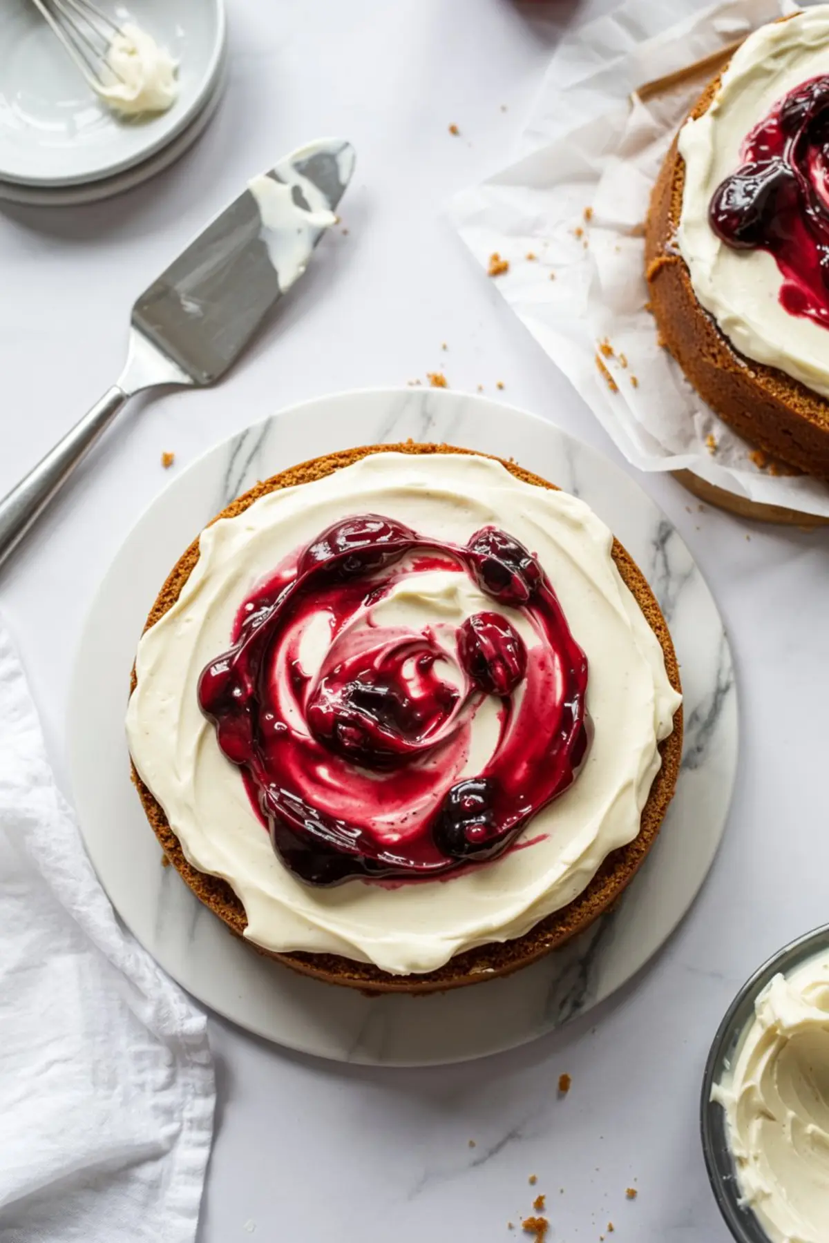 Top view of a round cake topped with smooth cream cheese frosting and a swirl of vibrant cherry or cranberry compote, placed on a marble serving board with baking tools nearby.
