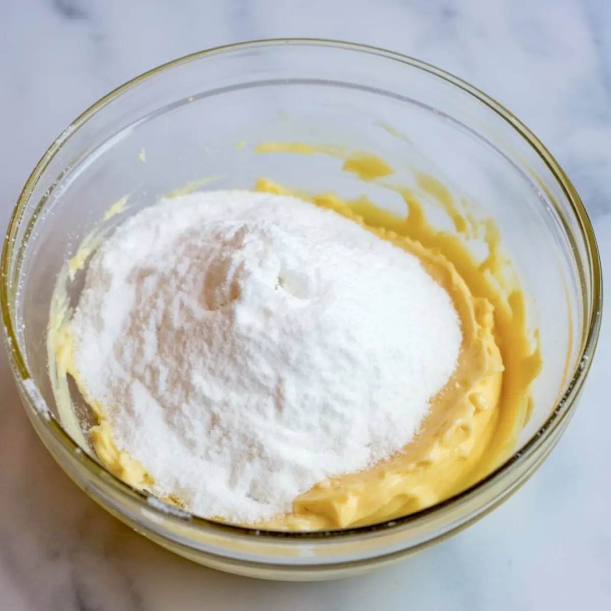 Powdered sugar piled on top of creamed butter mixture in a glass bowl, ready to be mixed into frosting.