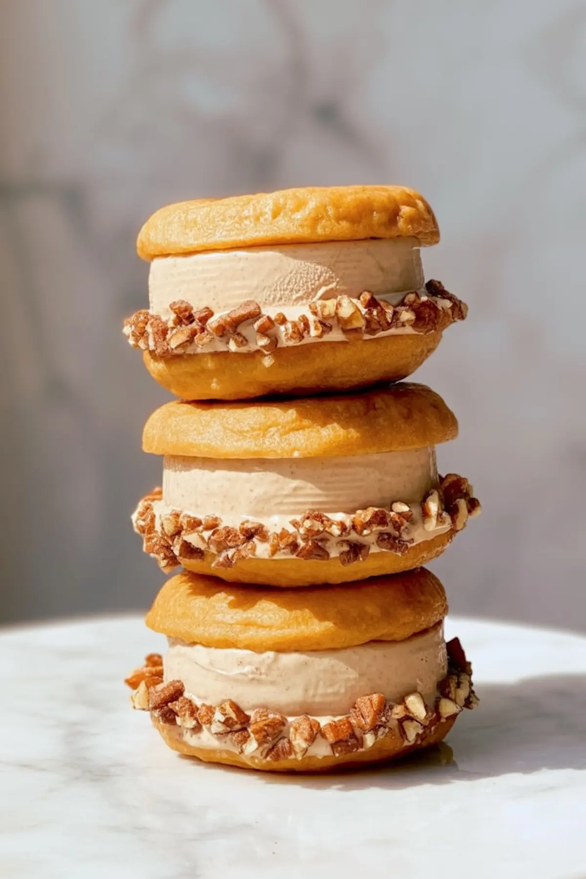 Three apple cider donut ice cream sandwiches stacked in a tower, with thick ice cream filling and edges rolled in chopped pecans, set against soft natural light.
