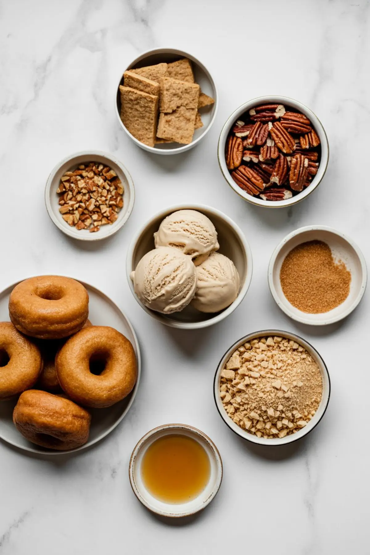 Flat-lay of ingredients for apple cider donut ice cream sandwiches, including donuts, vanilla ice cream, pecans, graham crackers, brown sugar, chopped nuts, and maple syrup arranged in small bowls.
