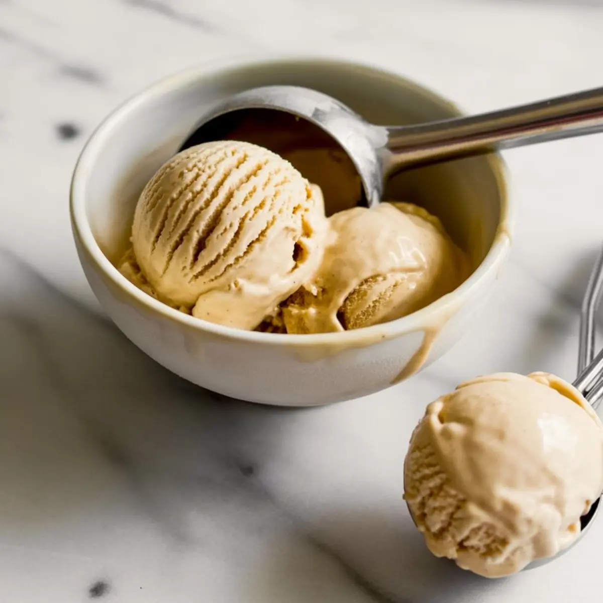 Vanilla ice cream scooped into a white bowl with an ice cream scooper resting on the edge, captured on a marble countertop in soft lighting.
