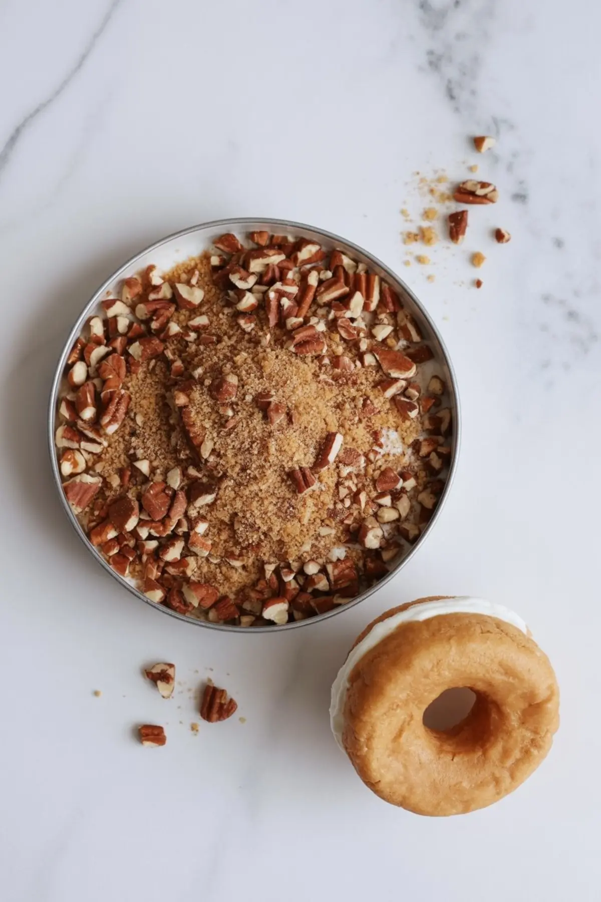 Bowl of brown sugar mixed with chopped pecans for coating ice cream edges, with a partially assembled apple cider donut ice cream sandwich nearby on a white surface.