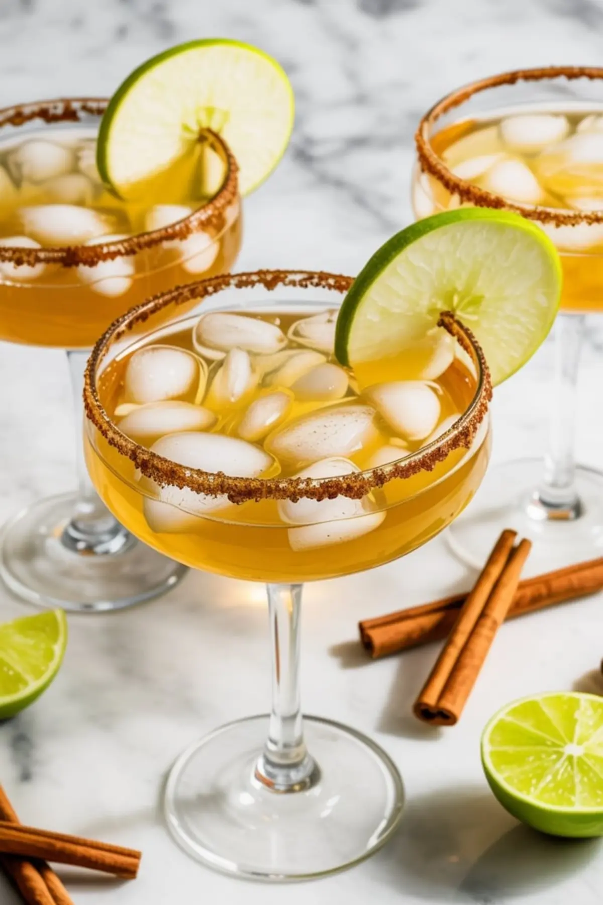 Three margarita glasses filled with apple cider margaritas over ice, garnished with lime wheels and rimmed with cinnamon sugar, arranged on a marble background with cinnamon sticks and halved limes.