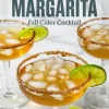 Close-up of apple cider margaritas in wide-rimmed cocktail glasses with ice, cinnamon-sugar rims, and lime slices, placed on a white marble table with fall-themed garnish.