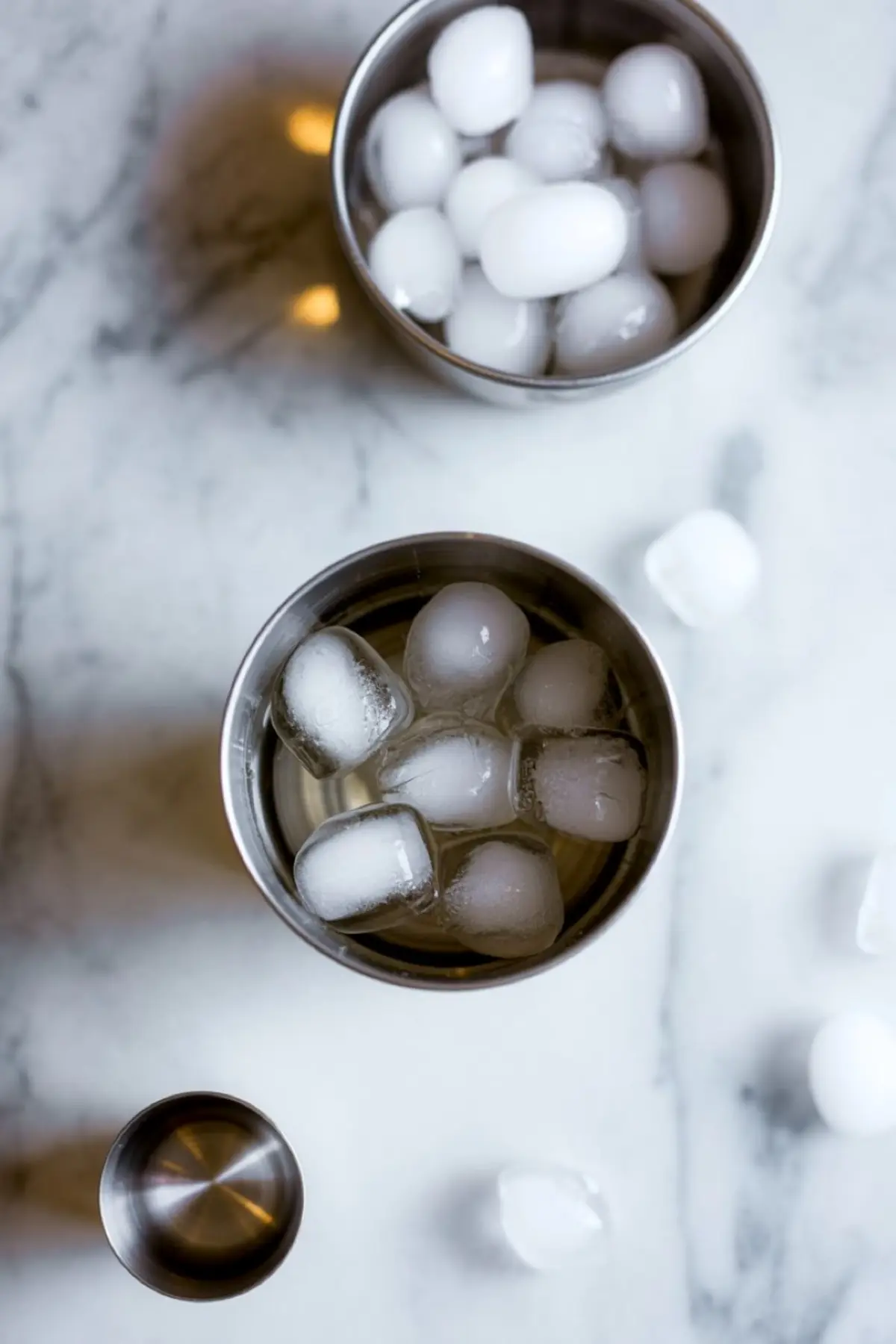 Two metal cocktail shakers filled with ice cubes on a white marble surface, accompanied by scattered ice and a small jigger, ready for making a chilled beverage.