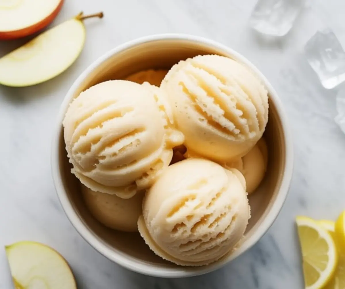Scoops of apple sorbet arranged in a white bowl with crisp texture and creamy color, surrounded by apple slices and ice cubes on a marble surface.
